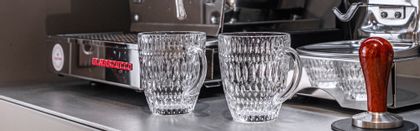 Two empty NACHTMANN Ethno hot beverage mugs in front of a portafilter machine and a coffee grinder. On the machine more Ethno coffee mugs are heeating up. To the right a coffee tamper.<br/>