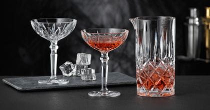 Le pichet NACHTMANN Noblesse rempli d'une boisson rouge et de glaçons et deux verres à cocktail NACHTMANN Noblesse, dont l'un est rempli de la même boisson rouge. À l'arrière-plan, le verre à cocktail vide et les glaçons.<br/>