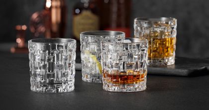 Quatre verres NACHTMANN Bossa Nova SOF, trois d'entre eux remplis de whisky ou d'autres boissons, l'un d'entre eux étant vide. En arrière-plan, des bouteilles d'alcool.<br/>