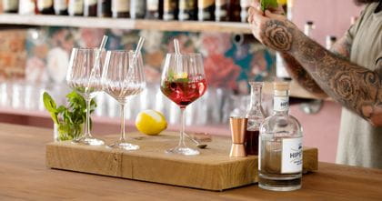 Verres à Gin et Tonic NACHTMANN avec pailles en verre sur un comptoir de bar. L'un d'eux est rempli d'un cocktail de gin rouge avec de la menthe et un zeste de citron. Derrière les verres se trouvent un citron et un verre avec des brins de menthe. À droite, les mains du barman et l'équipement du bar.<br/>