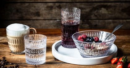 The NACHTMANN Ethno series on a wooden table. The hot beverage mug is filled with cappuccino, the tumbler with mineral water. The longdrink glass, filled with berry juice and the bowl, filled with a crunchy berry muesly, are standing on a white serving tray.<br/>