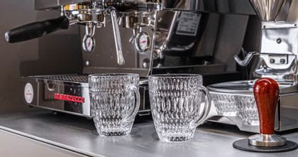 Two empty NACHTMANN Ethno hot beverage mugs in front of a portafilter machine and a coffee grinder. On the machine more Ethno coffee mugs are heeating up. To the right a coffee tamper.<br/>