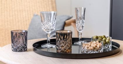 The NACHTMANN Noblesse collection on a serving tray. The smoke colored tumbler next to two Noblesse bowls, one filled with cashew nuts, one with olives. In the background are the Nobless wine glass and the Champagne glass.<br/>