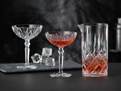 Le pichet NACHTMANN Noblesse rempli d'une boisson rouge et de glaçons et deux verres à cocktail NACHTMANN Noblesse, dont l'un est rempli de la même boisson rouge. À l'arrière-plan, le verre à cocktail vide et les glaçons.<br/>