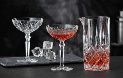 Le pichet NACHTMANN Noblesse rempli d'une boisson rouge et de glaçons et deux verres à cocktail NACHTMANN Noblesse, dont l'un est rempli de la même boisson rouge. À l'arrière-plan, le verre à cocktail vide et les glaçons.<br/>