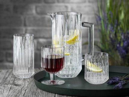 La série de verres en cristal NACHTMANN Jules sur un plateau de service rond. Le pichet et le gobelet sont remplis d'eau, de glaçons et de tranches de citron, à leur droite se trouve de la lavande. Le gobelet est rempli de vin rouge. Le verre à long drink à l'arrière-plan est vide.<br/>