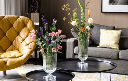 L'impressionnant vase Minverva de NACHTMANN, rempli de bouquets de fleurs, est posé sur des tables d'appoint rondes dans un salon. La pièce comprend un canapé, une lampe, un fauteuil en cuir et un tapis à motifs jaunes.<br/>