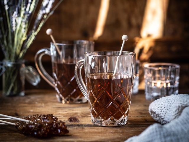 The NACHTMANN Noblesse tea mug with hot tea in it and rock candy. On the wooden table, there are more sticks with brown rock candy, a jar with lavender flowers, a second filled crystal tea mug and a shining tea light.<br/>