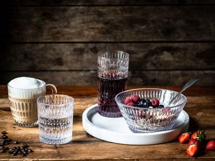 La série Ethno de NACHTMANN sur une table en bois. La tasse à boisson chaude est remplie de cappuccino, le gobelet d'eau minérale. Le verre à long drink, rempli de jus de baies, et le bol, rempli d'un muesly de baies croquantes, sont posés sur un plateau de service blanc.<br/>
