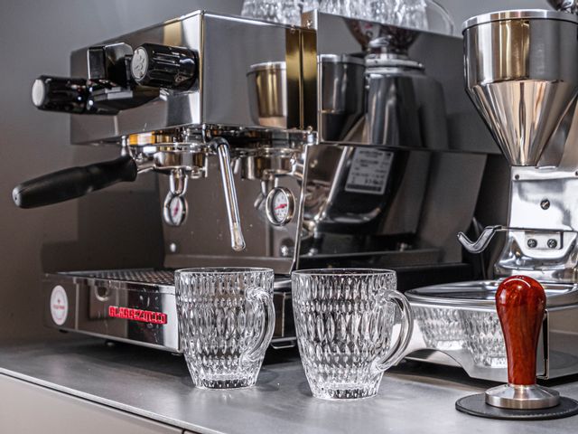 Two empty NACHTMANN Ethno hot beverage mugs in front of a portafilter machine and a coffee grinder. On the machine more Ethno coffee mugs are heeating up. To the right a coffee tamper.<br/>