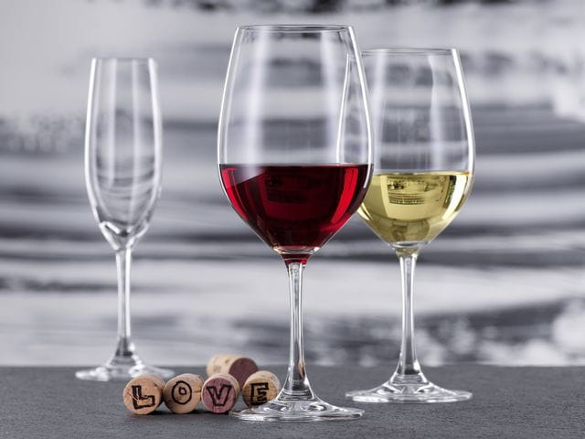 A with red wine filled Bordeaux glass and a with white wine filled white wine glass of the SPIEGELAU Winelovers collection. In the background, there is an empty Champagne flute. Five corks lie behind the Bordeaux glass, four of them showing letters of the word LOVE.<br/>