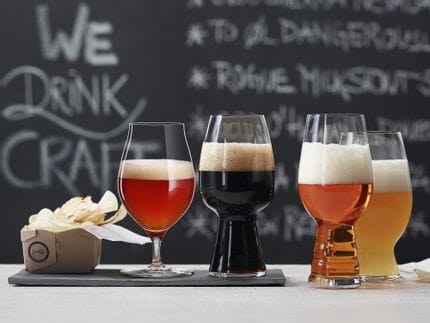 The with beer filled SPIEGELAU Craft Beer Glasses on a table in front of a blackboard with a menu. The glass for Barrel Aged beer and the Stout glass are standing on a slate plate, the IPA glass and the American Wheat Beer glass next to it on the table.<br/>