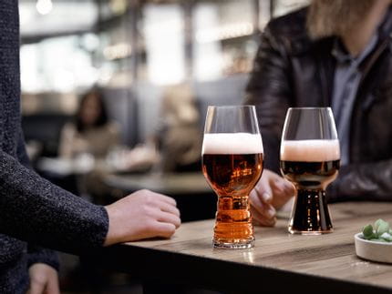 The SPIEGELAU Craft Beer Glasses IPA glass filled with IPA beer and the Stout glass filled with Stout beer standing on a wooden table. Behind them a hand reaching for the Stout glass.<br/>