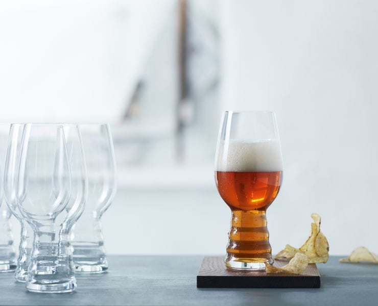 A group of SPIEGELAU Craft Beer Glasses for IPA on a table. One standing on a wooden coaster, filled with IPA beer. Next to this glass are some potato chips.<br/>