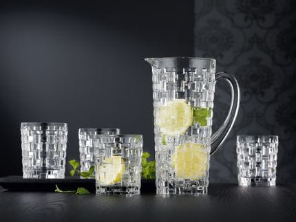 Le pichet Bossa Nova de NACHTMANN et quatre gobelets sur une table en bois noir. La carafe et l'un des gobelets sont remplis d'eau, de glaçons, de tranches de citron et de brins de menthe.<br/>