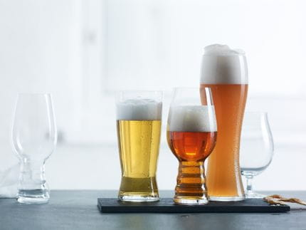 Four SPIEGELAU Beer Classics glasses in a row one is empty and the rest filled with different beers. The three filled glasses are on a black serving platter against a light background.<br/>