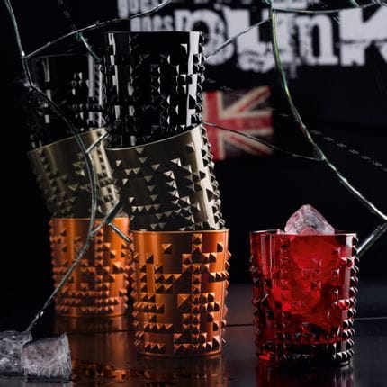 Quatre gobelets NACHTMANN Punk en noir de jais, cuivre et métal armé empilés devant un miroir brisé. À côté, le gobelet Punk en rubis est rempli de glaçons. Dans le miroir, il y a un Junion Jack et la ligne nextgen goes punk.<br/>