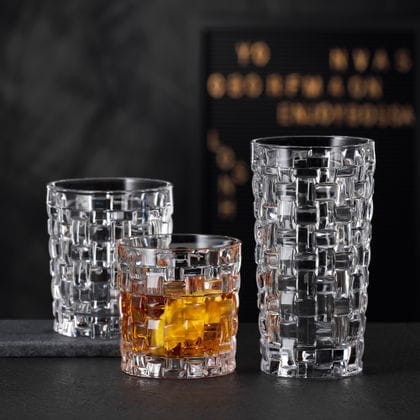 Le NACHTMANN Bossa Nova SOF rempli de whisky à côté du verre à long drink Bossa Nova vide sur un buffet en pierre noire. Derrière eux, le gobelet Bossa Nova vide.<br/>