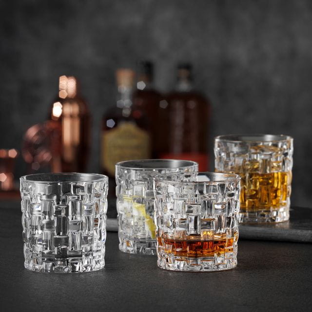 Four NACHTMANN Bossa Nova SOF glasses, three of them filled with either Whiskey or other drinks, one of them empty. In the background are liquor bottles.<br/>