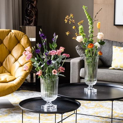 L'impressionnant vase Minverva de NACHTMANN, rempli de bouquets de fleurs, est posé sur des tables d'appoint rondes dans un salon. La pièce comprend un canapé, une lampe, un fauteuil en cuir et un tapis à motifs jaunes.<br/>