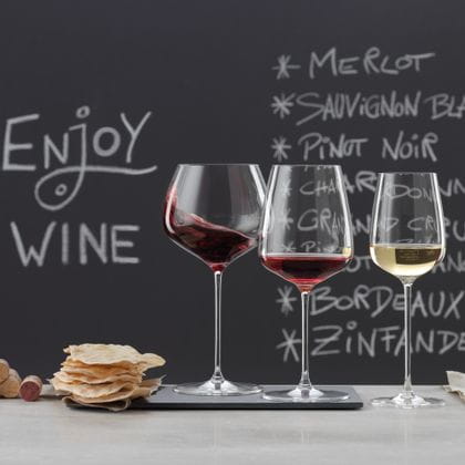 SPIEGELAU Willsberger Verres d'anniversaire sur un buffet en marbre. À gauche, un verre à bordeaux et un verre à vin blanc vides, au milieu une pile de bouchons et de chips, à droite un verre à bourgogne rempli, un verre à vin rouge rempli et un verre à vin blanc rempli. En arrière-plan, un tableau noir avec un menu et les mots "drink wine" (boire du vin).<br/>