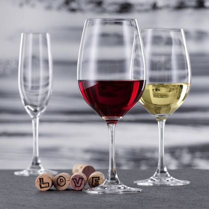 Un verre à bordeaux rempli de vin rouge et un verre à vin blanc rempli de vin blanc de la collection Winelovers de SPIEGELAU. À l'arrière-plan se trouve une flûte à champagne vide. Cinq bouchons de liège se trouvent derrière le verre de Bordeaux, quatre d'entre eux représentant les lettres du mot LOVE.<br/>