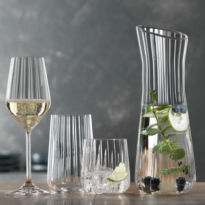 La série SPIEGELAU Lifestyle sur une table. Le verre à vin rouge rempli, le verre à vin blanc rempli, la coupe à champagne remplie, un verre à long drink vide, un gobelet rempli d'eau, de glaçons et d'une tranche de citron vert. À droite, la carafe contenant de l'eau infusée.<br/>