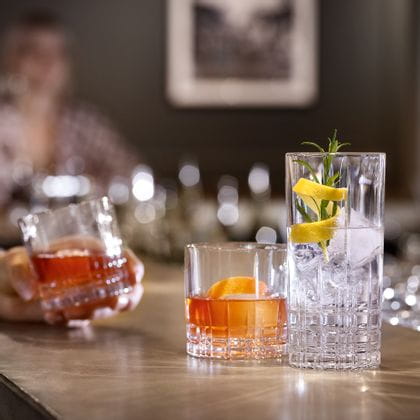 Un comptoir de bar avec deux verres SPIEGELAU Perfect Serve Collection. Le verre SOF est rempli d'une boisson orange et d'un zeste d'orange, le verre longdrink est rempli d'une boisson cocktail claire, de glaçons, de romarin et d'un zeste de citron. En arrière-plan, une main tient un autre verre SOF rempli.<br/>