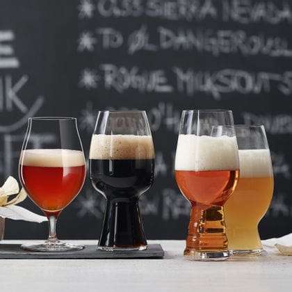 Les verres à bière artisanale SPIEGELAU remplis de bière sont posés sur une table devant un tableau noir avec un menu. Le verre à bière vieillie en fût et le verre à Stout sont posés sur une plaque d'ardoise, le verre à IPA et le verre à bière de blé américaine sont posés à côté sur la table.<br/>