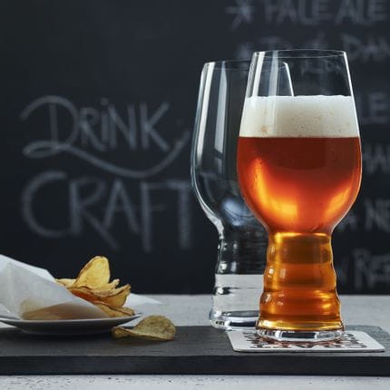 Two SPIEGELAU Craft Beer Glasses for IPA on a slate tray. One of them is filled with IPA beer, the glass behind is empty. Behind them is a plate with potato chips and a blackboard with a beer menu on it.<br/>