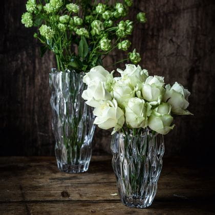 Le petit vase ovale NACHTMANN Quartz rempli de roses blanches devant le grand vase NACHTMANN Quartz rempli de fleurs blanches sur un buffet en bois foncé.<br/>