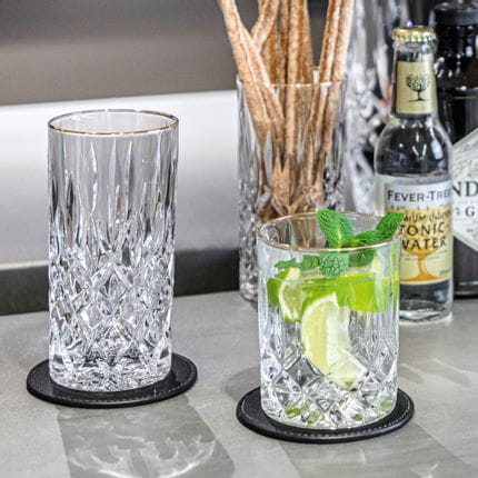 The NACHTMANN Noblesse Gold longdrink glass next to the tumbler filled with a gin and tonic cocktail. In the background one more longdrink glass filled with crissini and a bottle of tonic water.<br/>