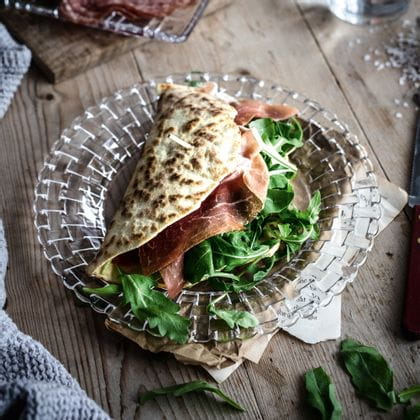 Der kleine NACHTMANN Bossa Nova Teller mit einem Wrap, gefüllt mit Schinken und Rucola, auf einem Holztisch.<br/>