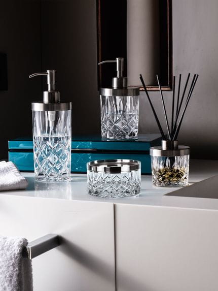 The NACHTMANN Spa Noblesse series on a bathroom sink in front of a mirror. The scent diffuser is filled with fragrance and black sticks. The small and the tall soap dispenser are filled with clear liquid. The storage jar is empty.<br/>
