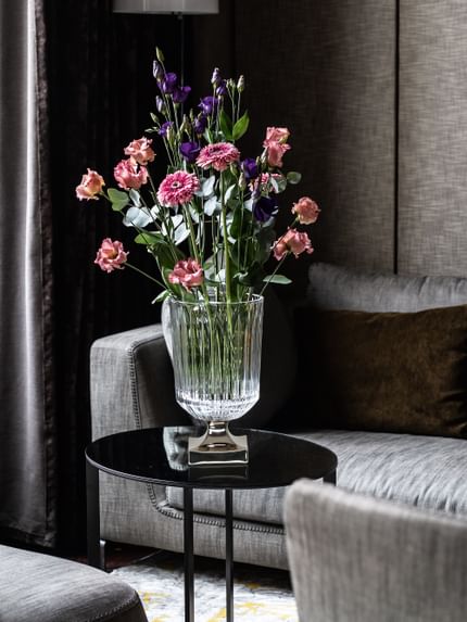 The NACHTMANN vase Minerva with a platinum coated foot and a large bouquet of flowers on a round side table. In the background is a grey couch with a brown pillow.<br/>