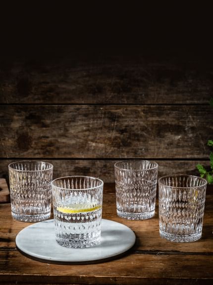 Four NACHTMANN Ethno tumblers on a wooden sideboard. One of them is filled with water and a slice of lemon and stands on a round stone platter. Around the tumblers are lemons, slices of lemon, and mint sprigs.<br/>