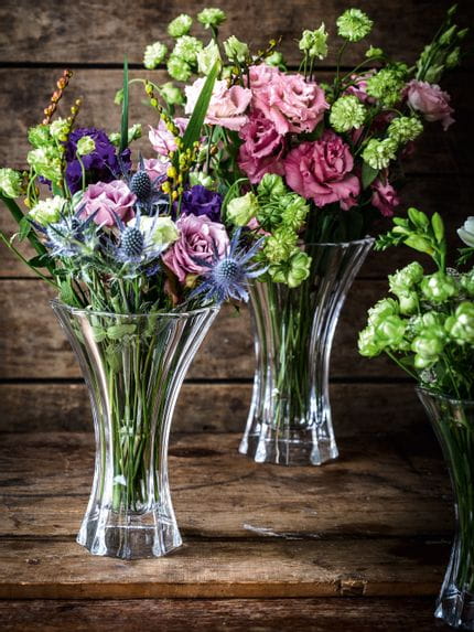 Die Kristallvasen NACHTMANN Saphir, gefüllt mit bunten Blumensträußen in Rot, Grün, Violett und Blau auf einem hölzernen Anrichte.<br/>