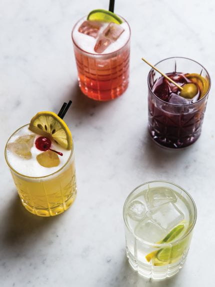 Four Nachtmann Highland Tumblers filled with red, yellow, purple and clear cocktails on marble bench top.