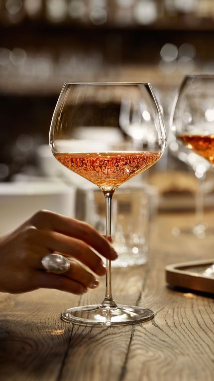 A hand holding the SPIEGELAU Burgundy glass of the series Willsberger Anniversary, filled with rosé colored sparkling wine. Further back on the wooden table, there are more glasses.<br/>